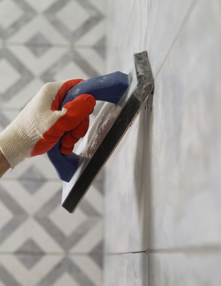 Close-up of a hand in a red glove using a blue grout float to apply grout on light grey tiles with subtle marble texture.